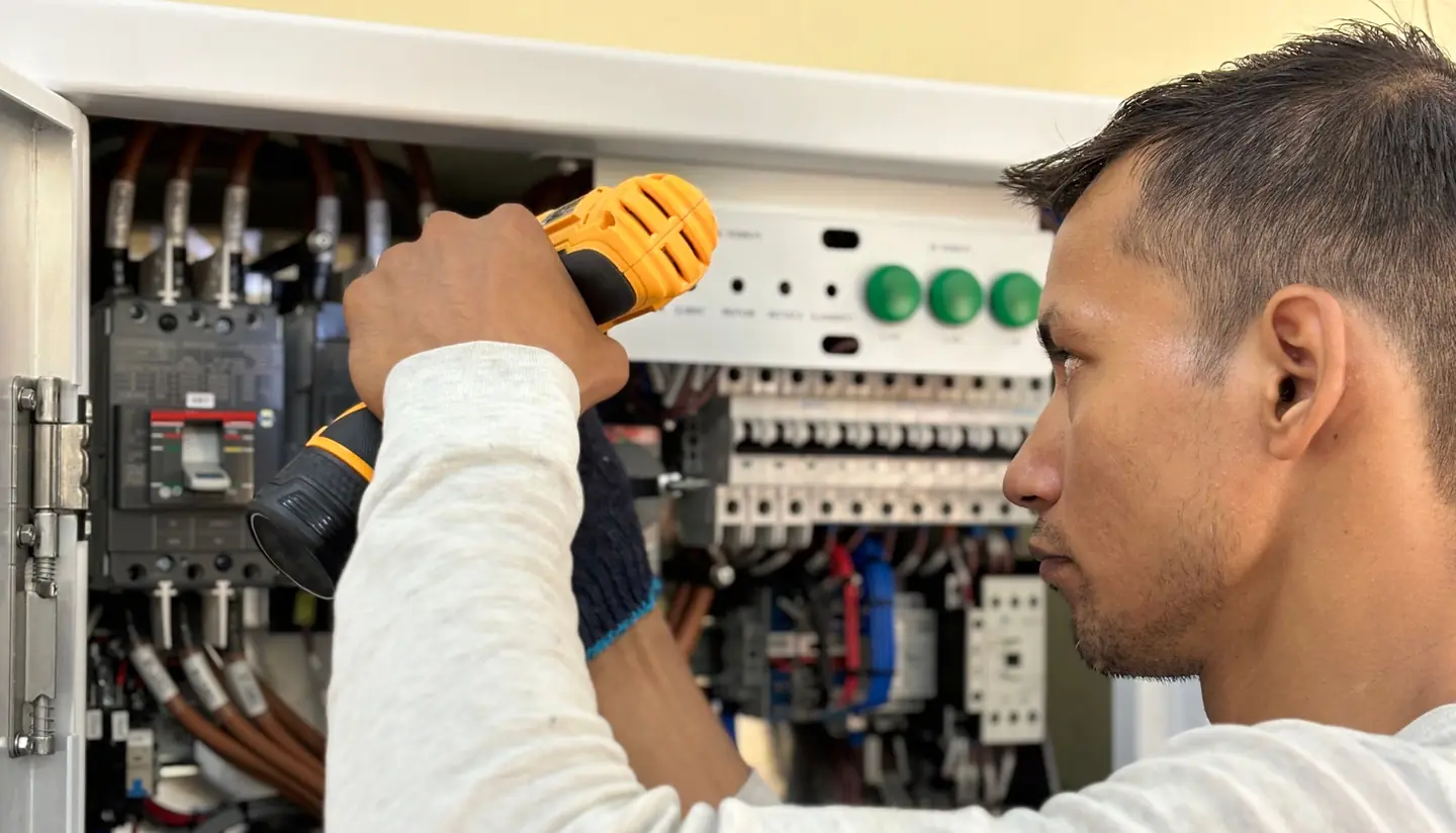 An electrician in a white top working on an open control panel with a cordless drill.